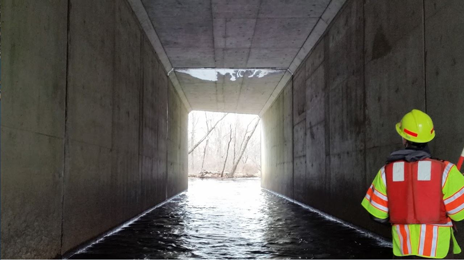 A bridge inspector wearing a safety vest, hard hat, and a life vest stands inside a tall, long, concrete box culvert, looking through the span to inspect the structure. There is water flowing through the culvert and the opening shines brightly at the end, revealing trees in the distance.
